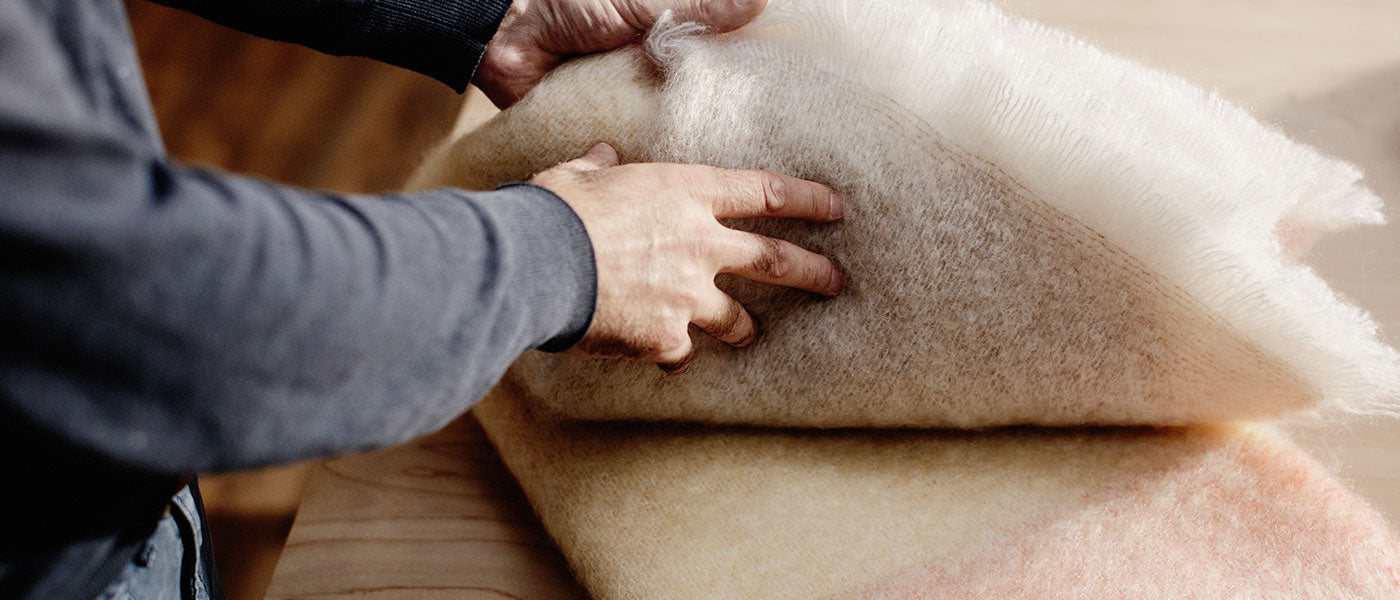 Hands with dark sleeves unfolding a pale beige and pink woollen blanket on a wooden table